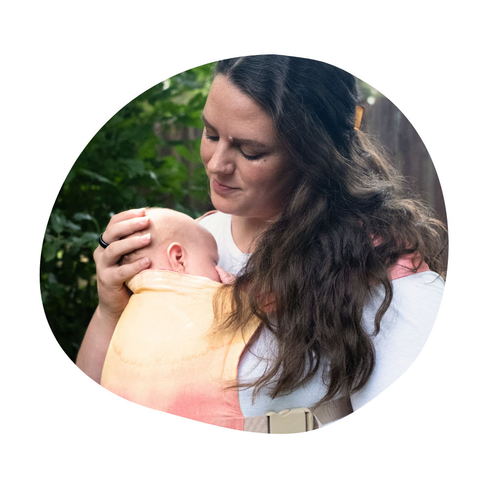 Aurora looks down tenderly at her sleeping baby Callum, held against her chest in a pink-and-yellow Vesper Sprout carrier, with green foliage behind her.