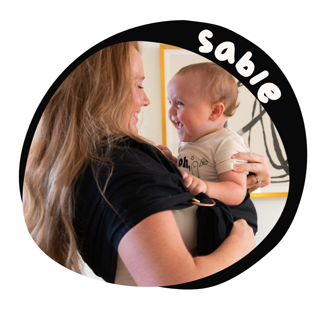 A woman with long blonde wavy hair holds a baby in a pure black ring sling. The mother and baby are grinning at each other, full of love.