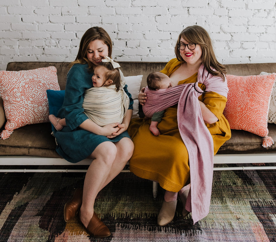 hope&plum founders Skye and Mallory sit together on a couch with their babies — Skye in a teal dress cradling a toddler in a cream striped wrap, Mallory in mustard yellow nursing a newborn in a dusty pink ring sling.