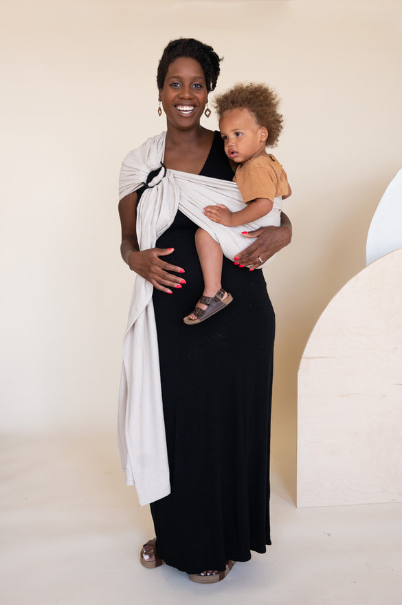 Pregnant Black mother holding her toddler in a gray ring sling on her hip. 