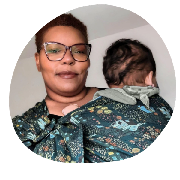 black woman with brown hair and glasses holding a baby in a green ring sling that is printed with houses and greenery