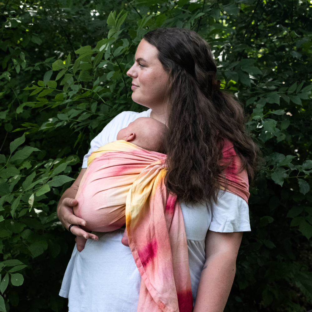 brunette woman wearing her infant in a pink and yellow watercolor sling with foliage in the background