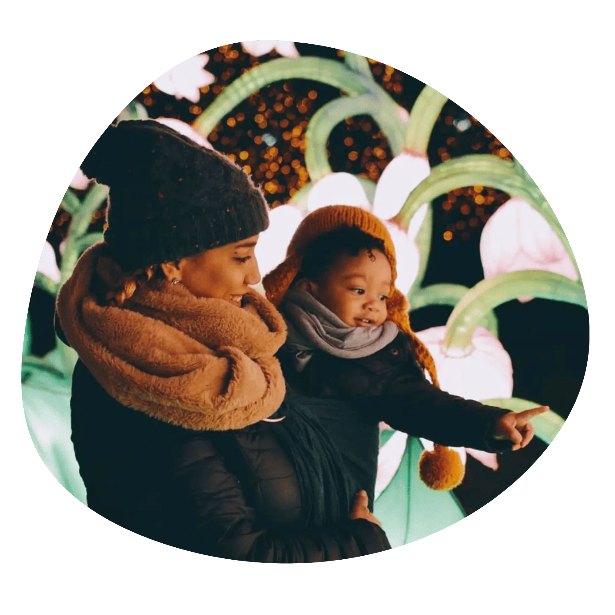 A brown-skinned woman bundled up for winter stands in profile as she wears her brown-skinned, black and curly-haired baby in a  hip carry in a ring sling. The baby is also bundled and pointing off frame.