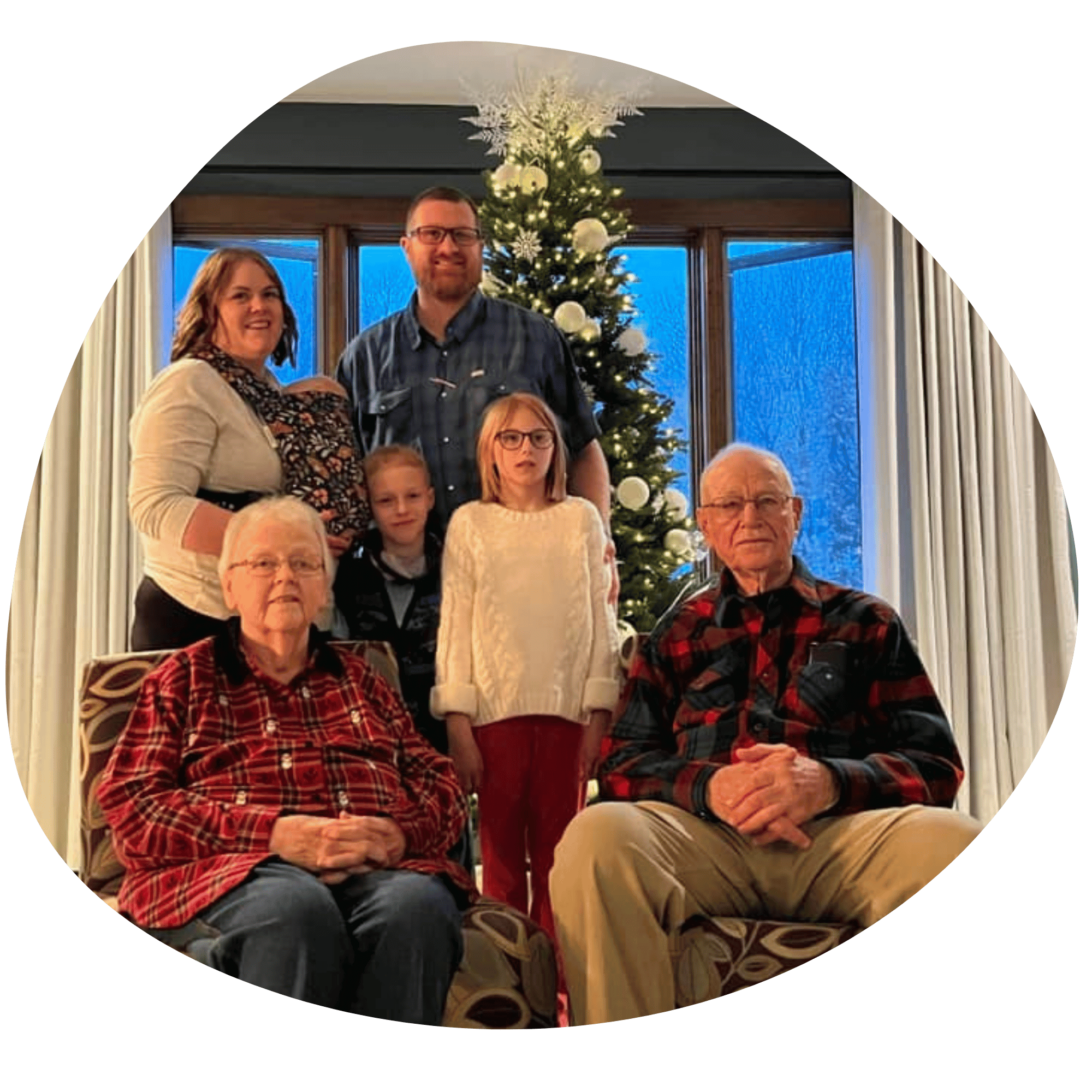 A family gathers in front of a Christmas tree for a posed picture. Two older family members sit in chairs in the front while a family of 5 - 2 adults and 3 children - stand behind. The mother wears the youngest child in a Lark on her front. 