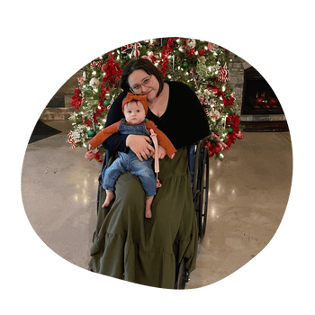 A light-skinned woman with shoulder length dark brown hair sits in a wheelchair in front of a Christmas tree. She embraces the baby she's holding as she smiles at the camera. The light-skinned baby sits on one of her legs and faces the camera.