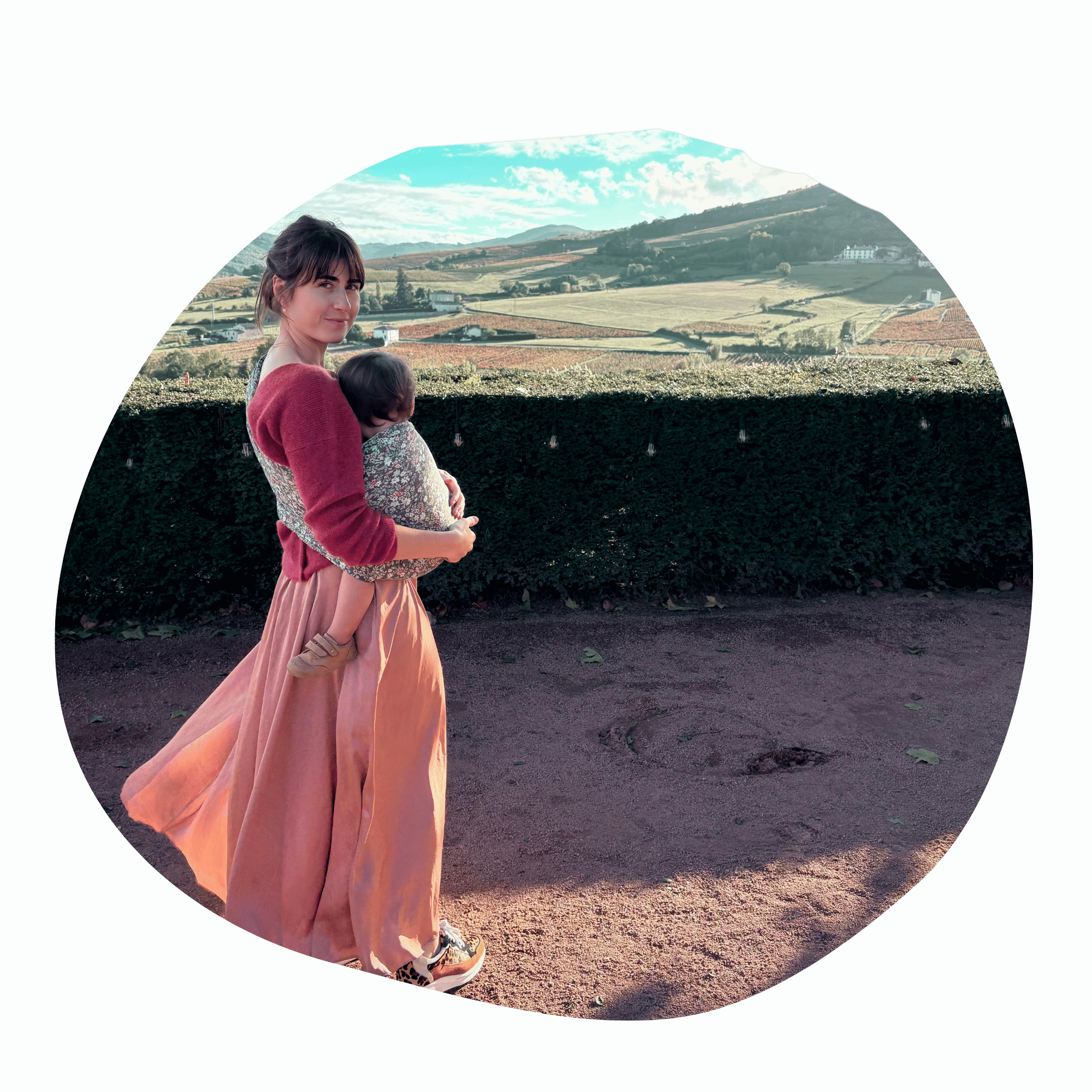 A light skinned brown haired straight sized woman stands outside and looks over her shoulder at the camera while she wear her light skinned brown haired baby  in a hip carry in a ring sling. She stands in front of bucolic landscape.