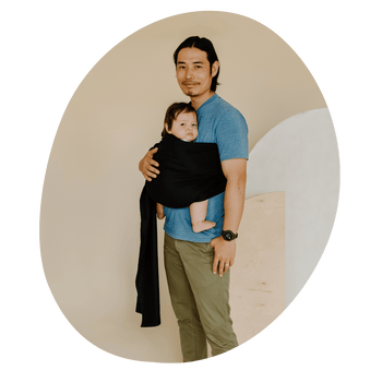 A BIPOC straight size man smiles at the camera while wearing his baby on his front in a ring sling.