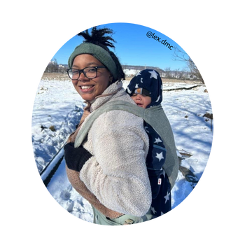 A brown skinned woman with black hair and glasses stands in profile and smiles at the camera. She is cold weather babywearing in a Lark.
