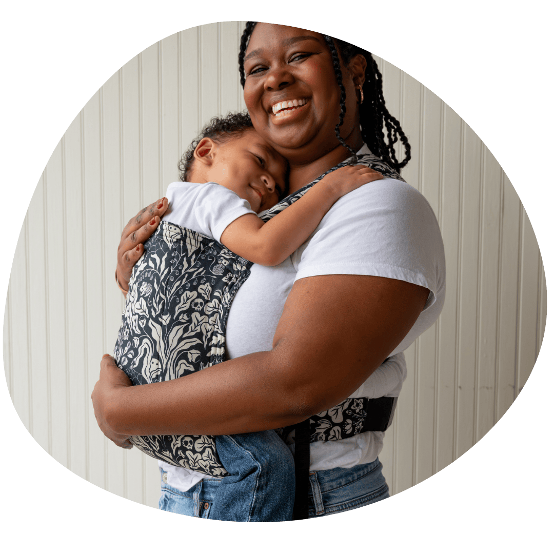 A woman smiles at the camera and stands in profile while wearing her older baby in a Lark on her front. Her baby appears to  be smiling and hugging her. 