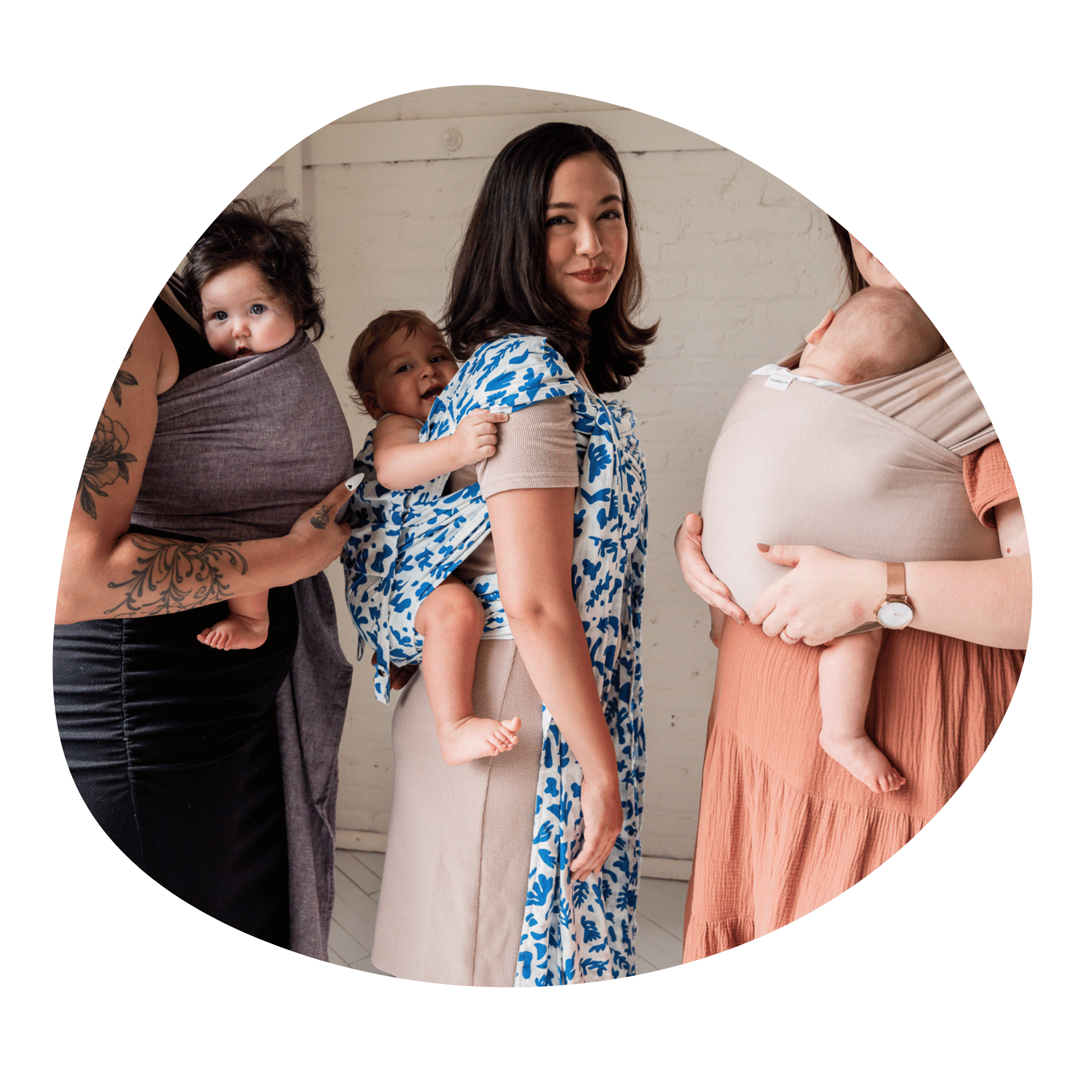 Three caregivers stand while carrying babies: one in a ring sling front carry, one in a back carry in a meh dai, and one in a stretchy wrap. The middle caregiver is the only one seen in full.