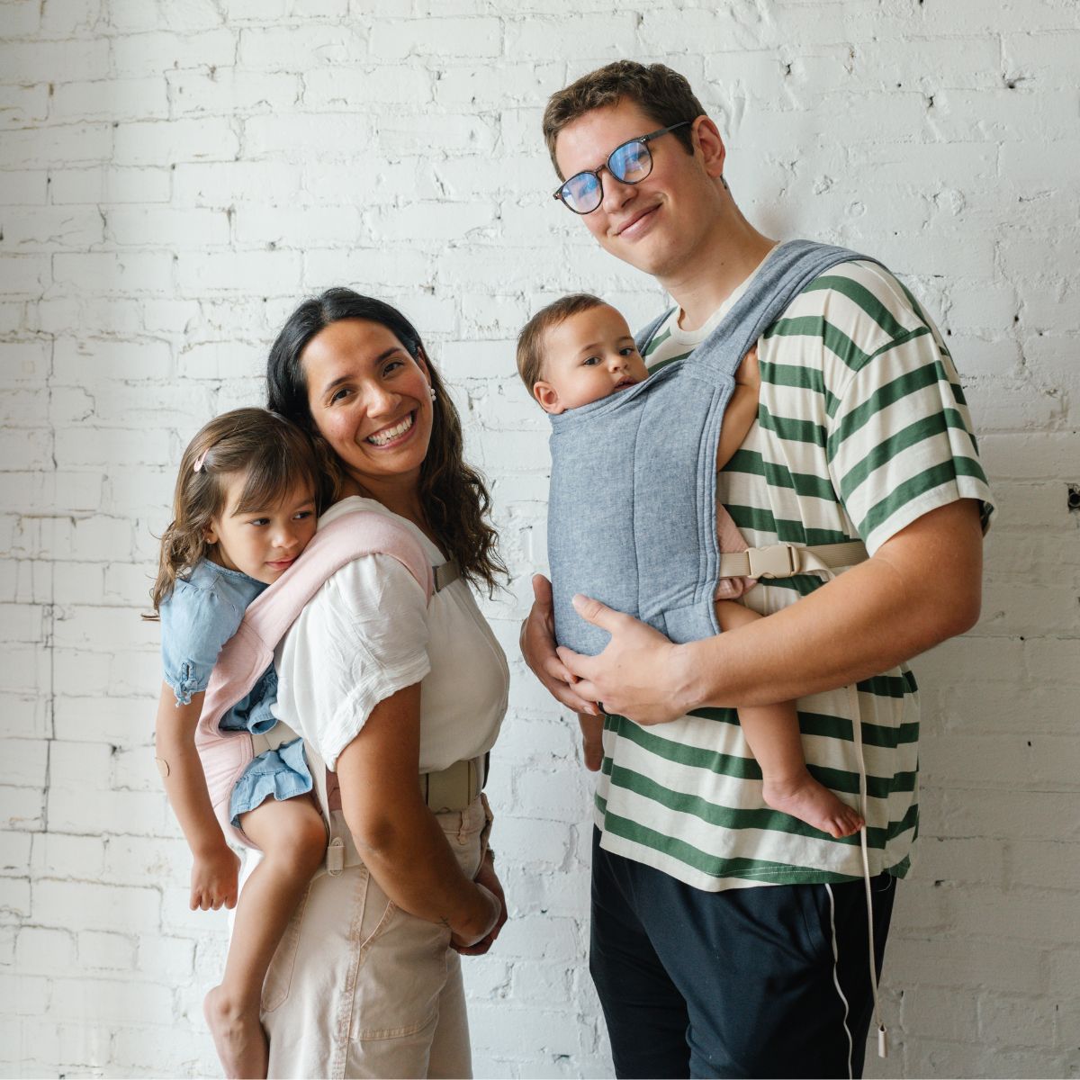A BIPOC mother wearing her baby on her back in a pink Lark Baby Carrier and a white father wearing an infant on his front in a blue Lark Baby Carrier.