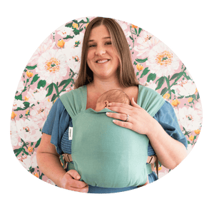 A white plus sized woman smiles at the camera while wearing her newborn in a hope&plum Sprout Newborn Carrier on her front