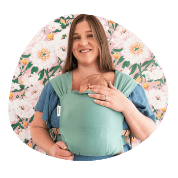 A white plus sized woman smiles at the camera while wearing her newborn in a hope&plum Sprout Newborn Carrier on her front