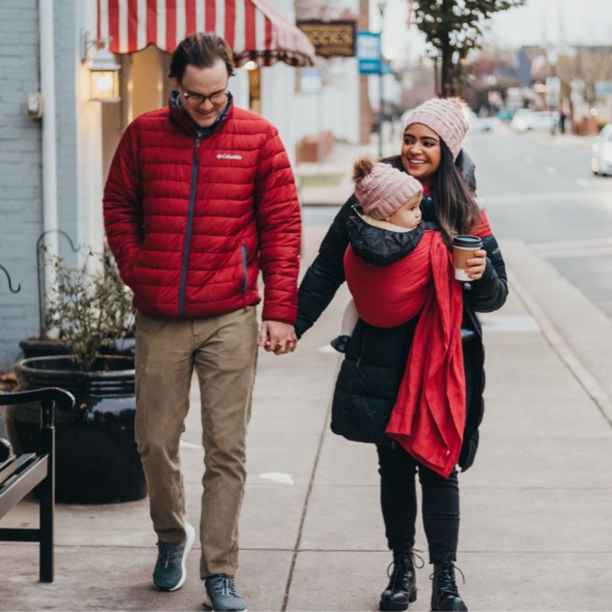A straight-size white man and a straight-size BIPOC woman walk hand -in-hand. They're both dressed for the cold. The woman wears a bundled baby on her front in a hope&plum Aster ring sling