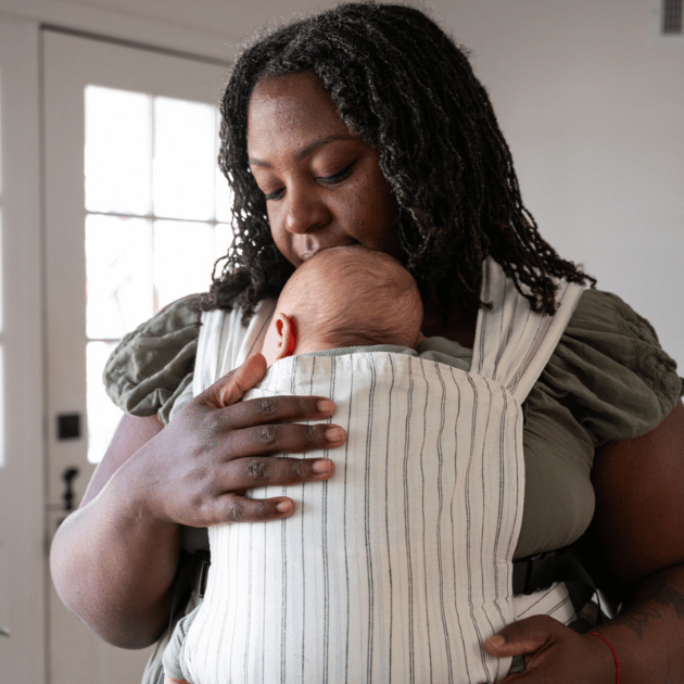 A BIPOC plus size woman looks down and embraces her infant worn on her front in a hope&plum Sprout Newborn Carrier