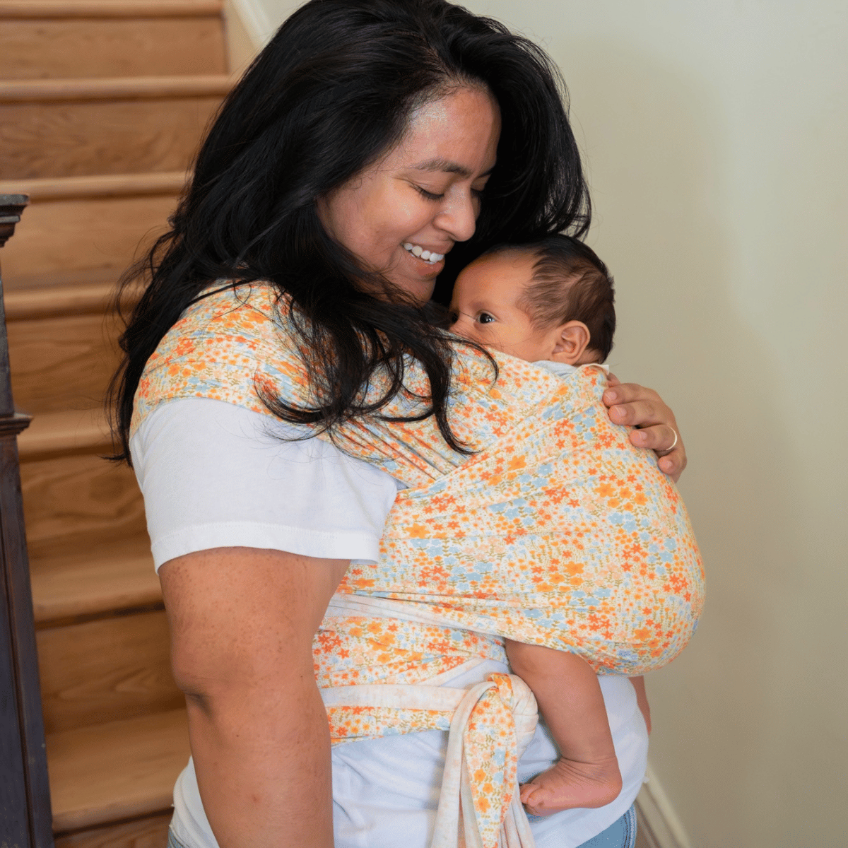 A straight size, BIPOC woman looks down and smiles at her baby, who is being carried in a stretchy wrap.