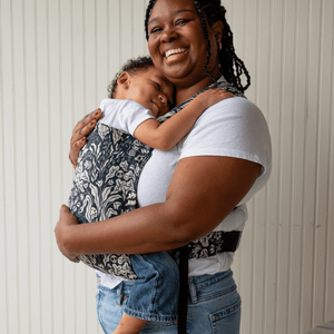 A mid size BIPOC woman smiles and wears her older baby in a hope&plum Lark on her front
