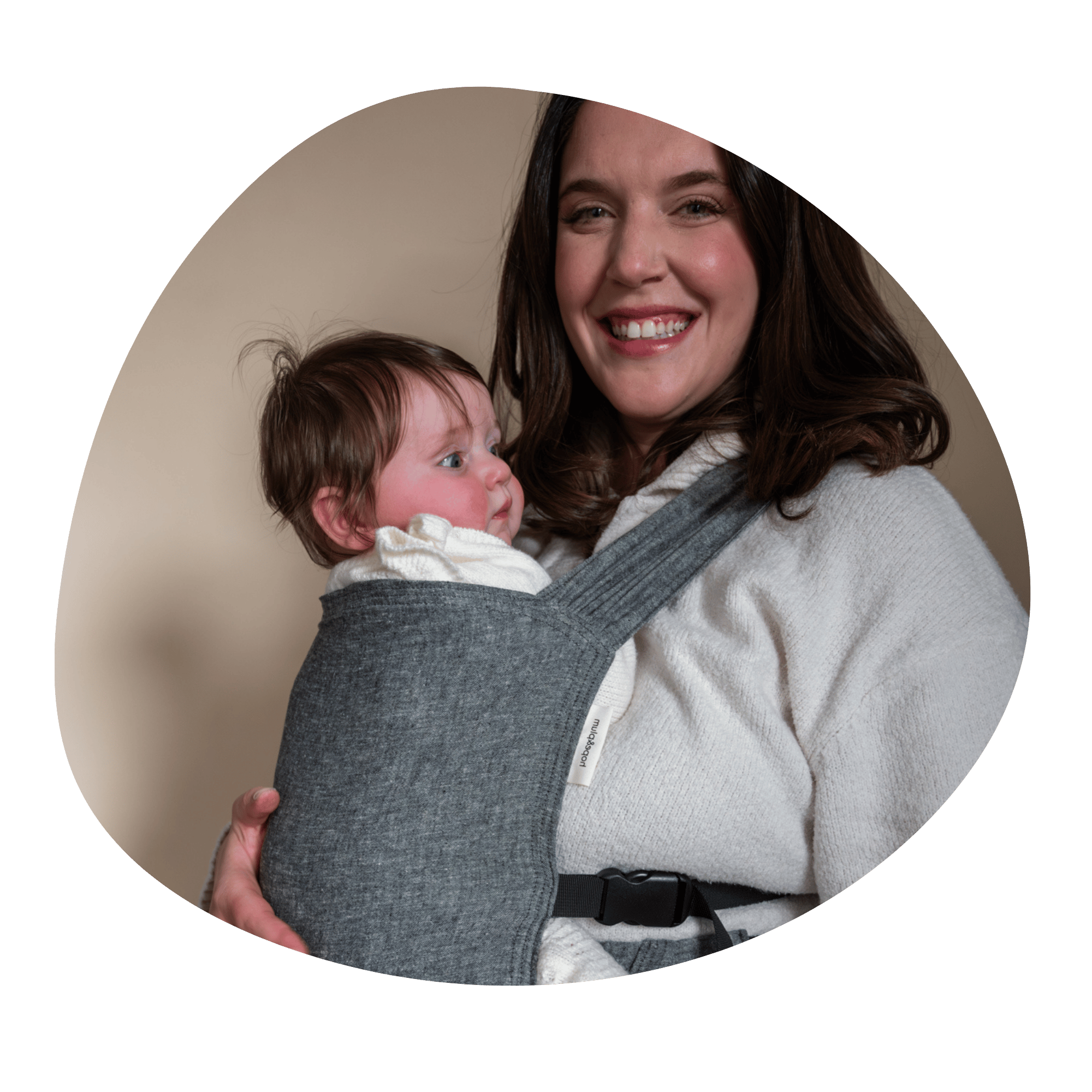 A white woman smiles at the camera while embracing her older infant worn on her front in a Sprout