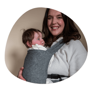 A white woman smiles at the camera while embracing her older infant worn on her front in a Sprout