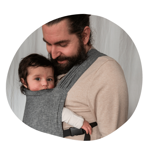 A white straight size bearded man smiles down at his infant worn on his front in a hope&plum Sprout Newborn Carrier