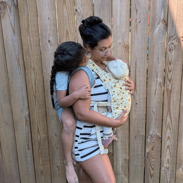 A straight size BIPOC woman looks down at a baby worn on her front in a hope&plum Baby Lark and wears a big kid on her back in a Kid Lark