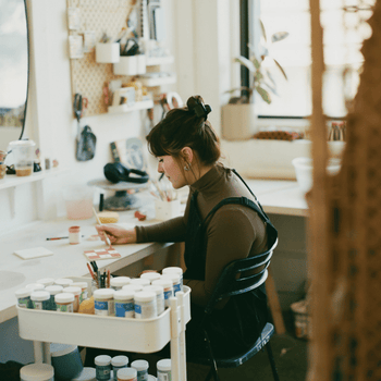 A straight size white woman works paints a tile in pink checker print and flowers in her bright indoor art studio