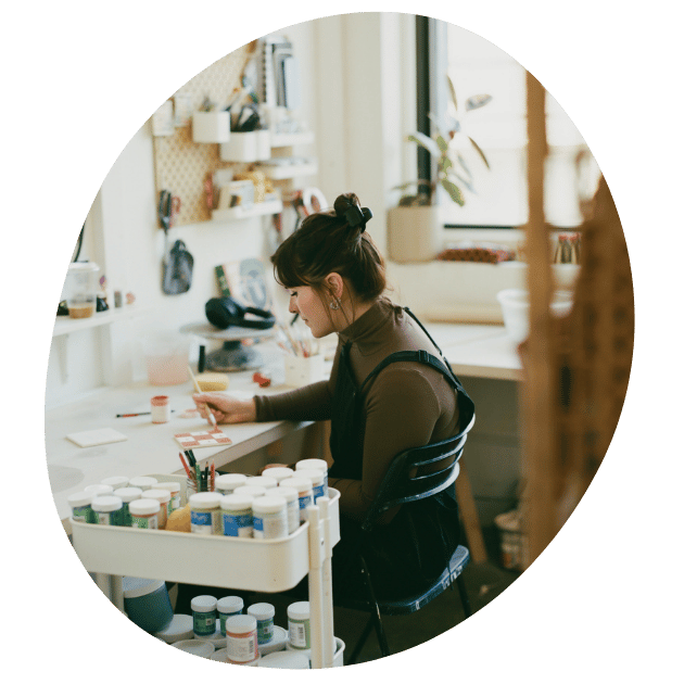 A straight size white woman works paints a tile in pink checker print and flowers in her bright indoor art studio