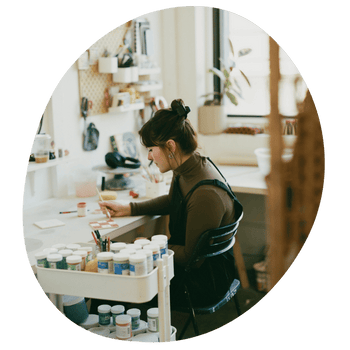 A straight size white woman works paints a tile in pink checker print and flowers in her bright indoor art studio