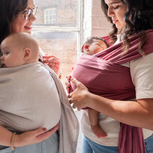 White mother wearing her baby in a gray ring sling talking to another mother wearing her newborn in a rose-colored baby wrap.