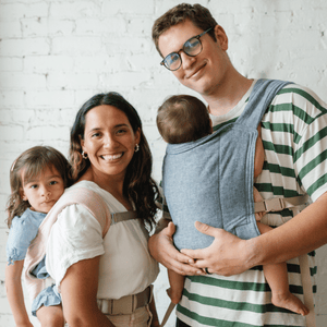 A family of four: A BIPOC woman carries their toddler daughter on her back, in a Lark. A white male carries their  baby on his front, in a lark.
