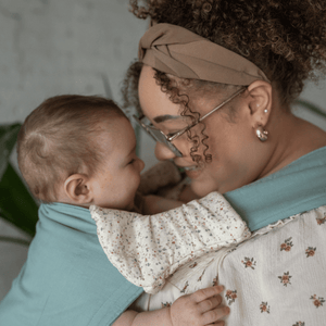 a BIPOC plus size mom smiles down at her baby in a carrier on her front. there is a pair of drool pads on the carrier straps.