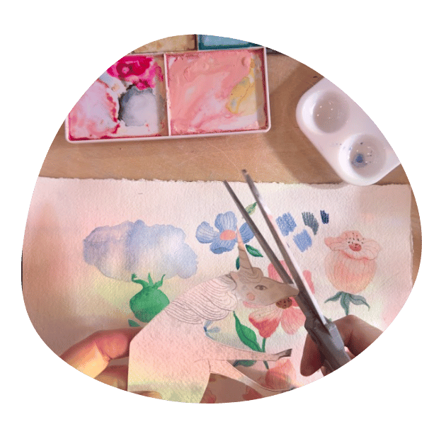 A close up on the design process for a print. A pair of white hands hold a paper unicorn and scissors above a watercolor painting of flowers