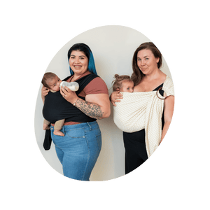 Two women stand in nearly profile. The first wears her baby in a stretchy wrap and bottle feeds. The second wears her baby in a ring sling and nurses.