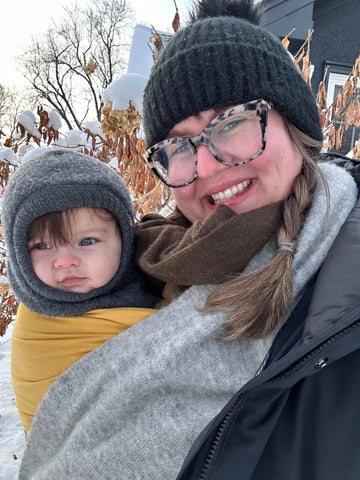 A white woman wearing a yellow ring sling with her baby in it. There is snow around them.