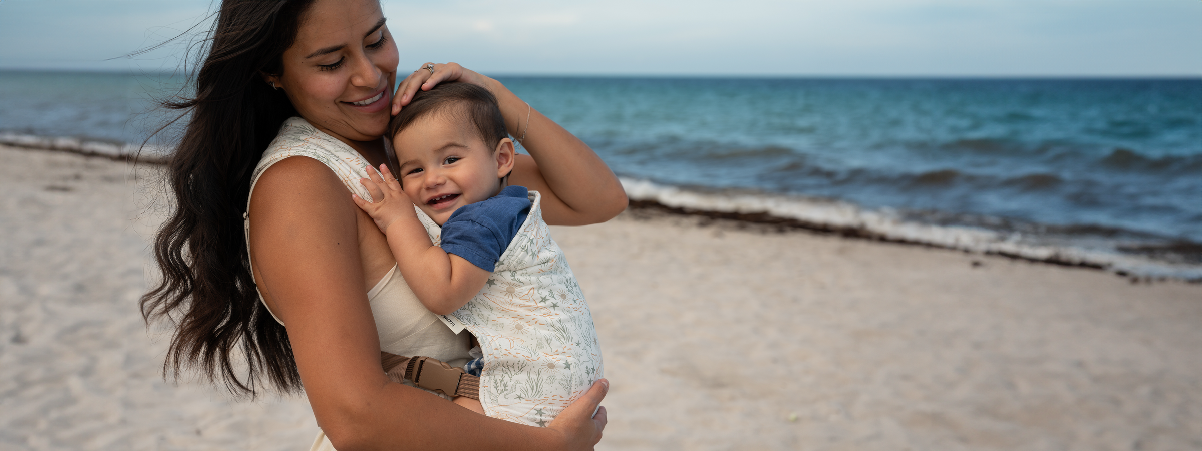 A smiling caregiver holds a grinning baby in the Cove Lark — a cream woven fabric with delicate sea creatures and coral in soft sage and terracotta — on a breezy sandy beach.