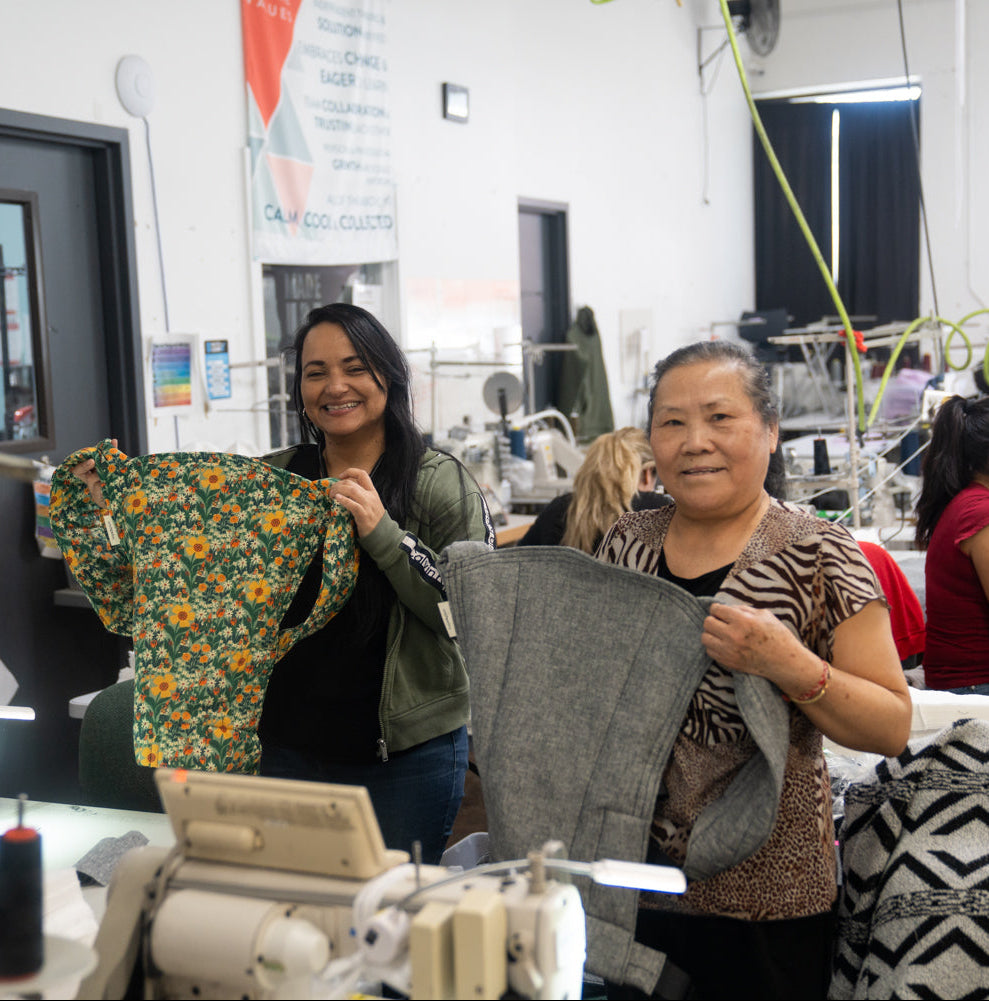 Two women holding up baby carriers sewn in Minnesota with a factory background.