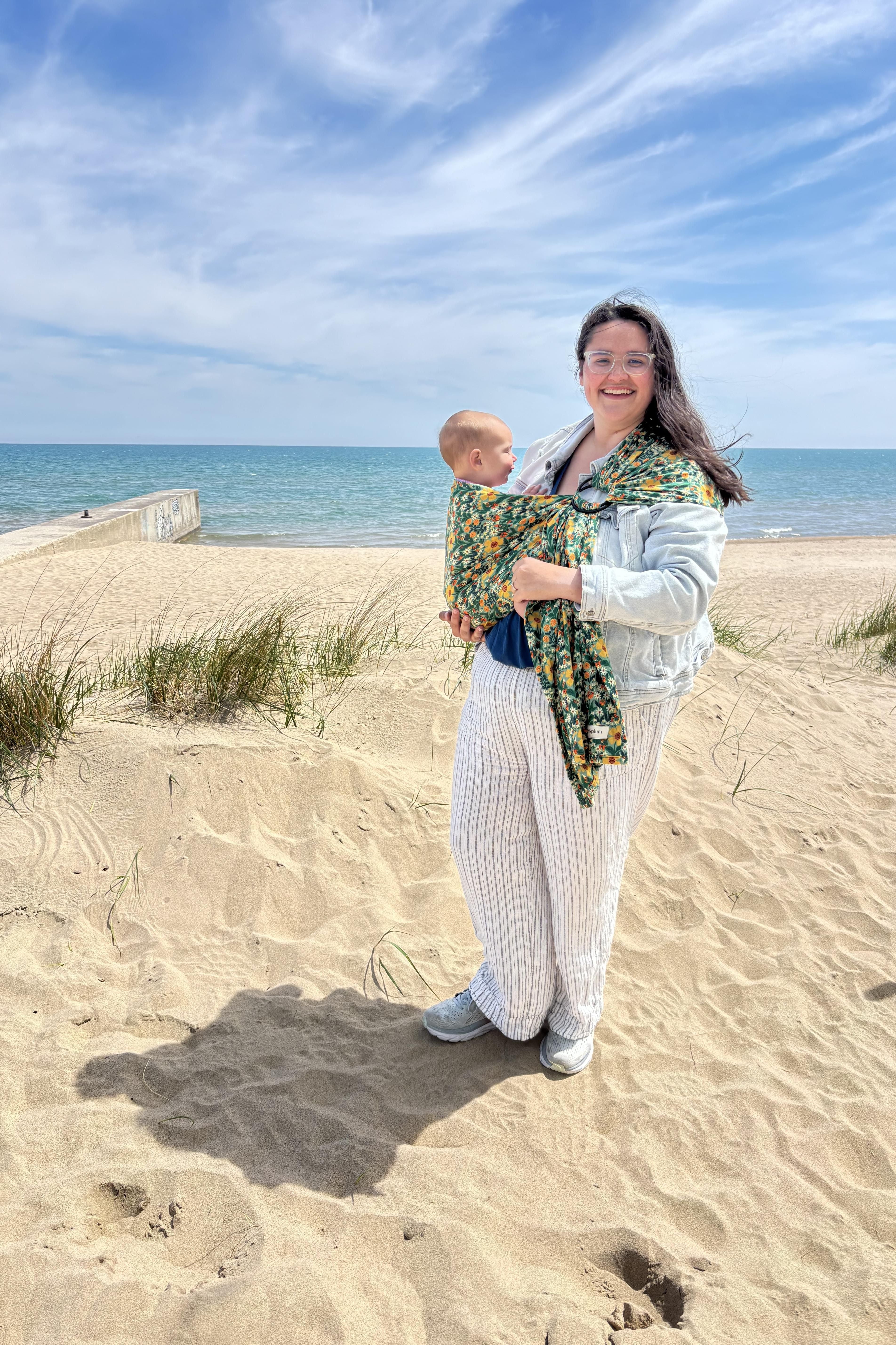 Woman holding a baby in a green floral ring sling on a sandy beach with ocean and sky in the background