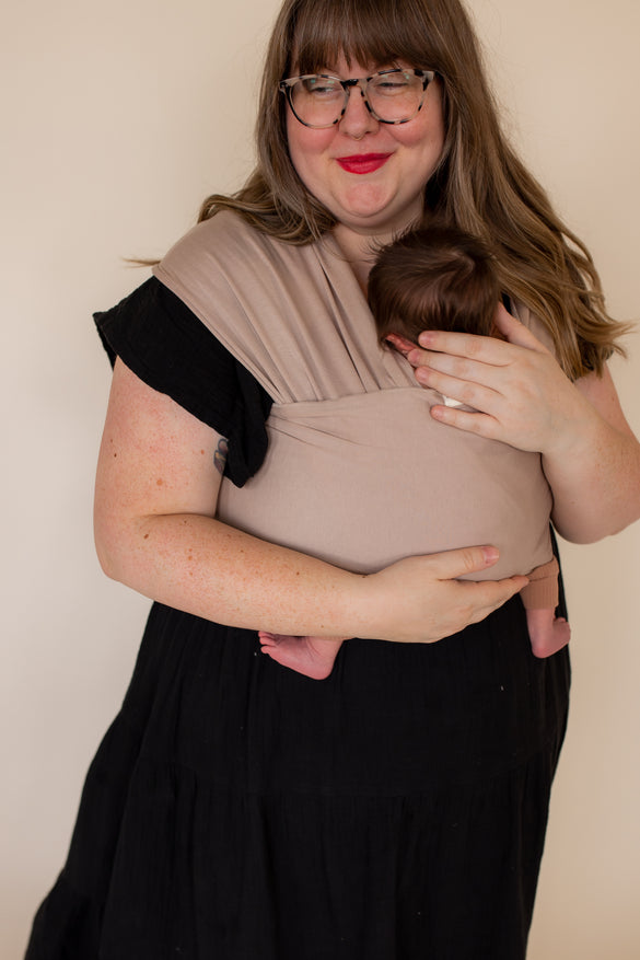 Plus-size mother wearing newborn in a tan-colored wrap baby carrier.