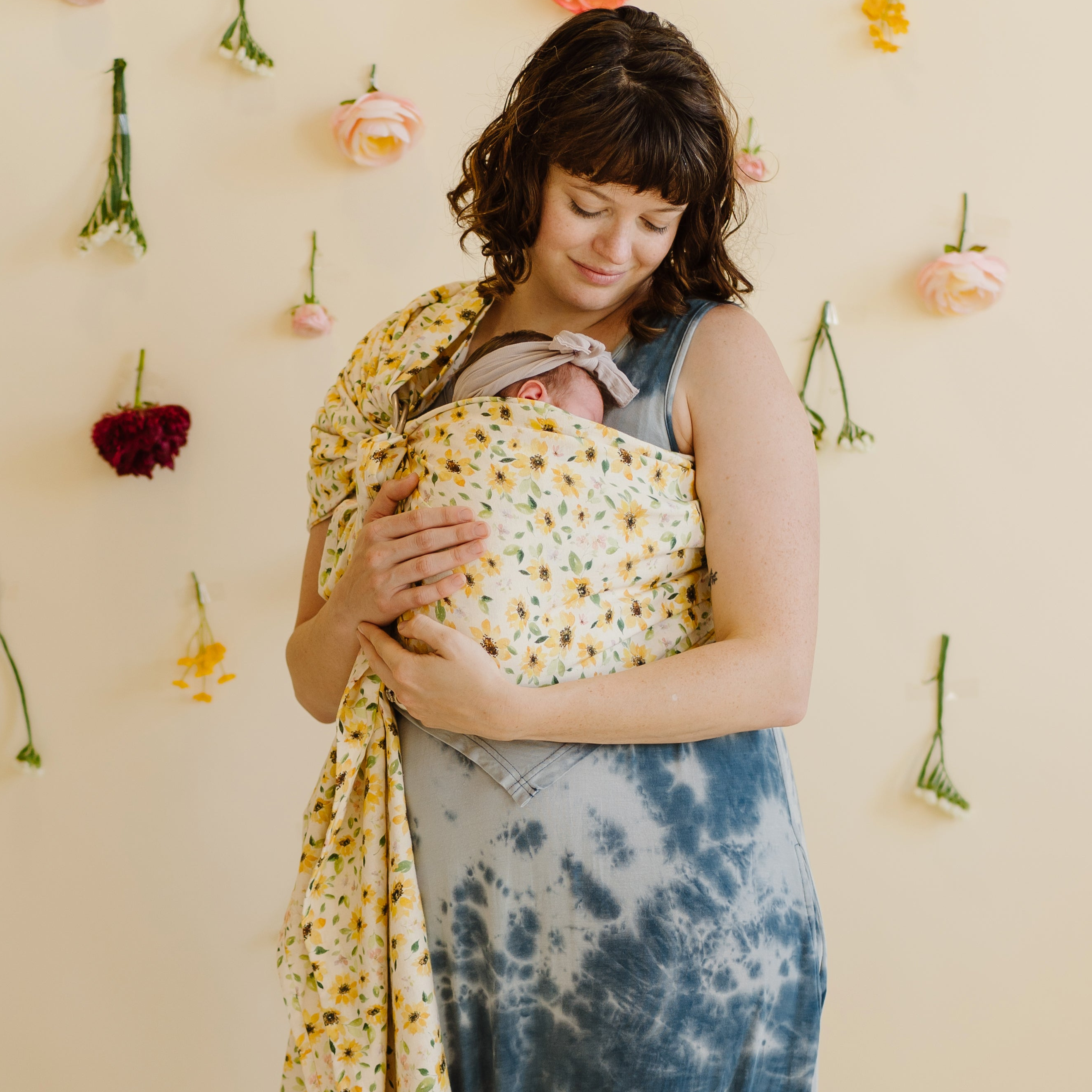 Woman holding a baby wrapped in a floral ring sling against a wall with hanging flowers.