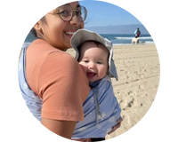 Mother holding smiling baby in blue baby carrier at the beach. 