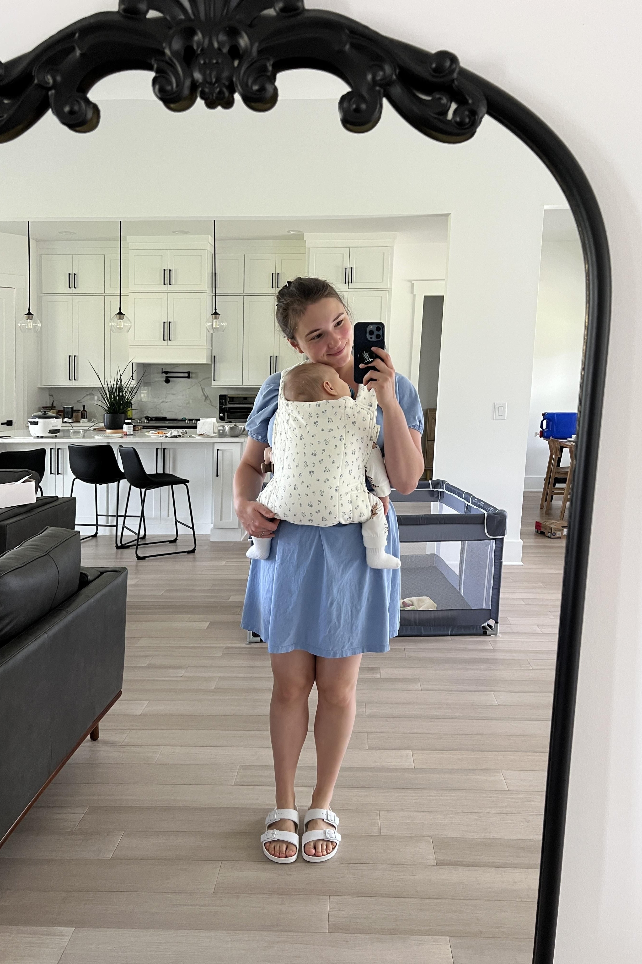 Person holding a baby in a baby carrier in a mirror selfie in a modern kitchen.