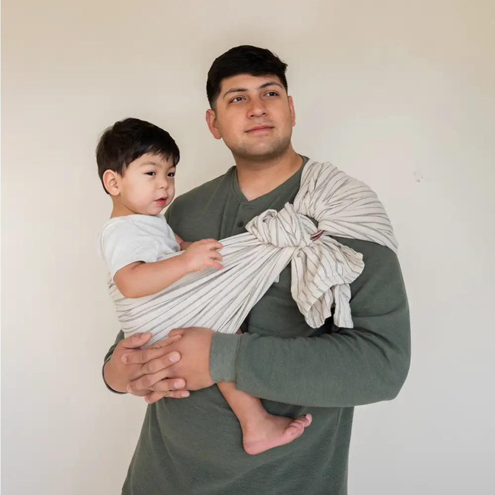 Father wearing white and black stripe ring sling baby carrier on his hip with his son. 