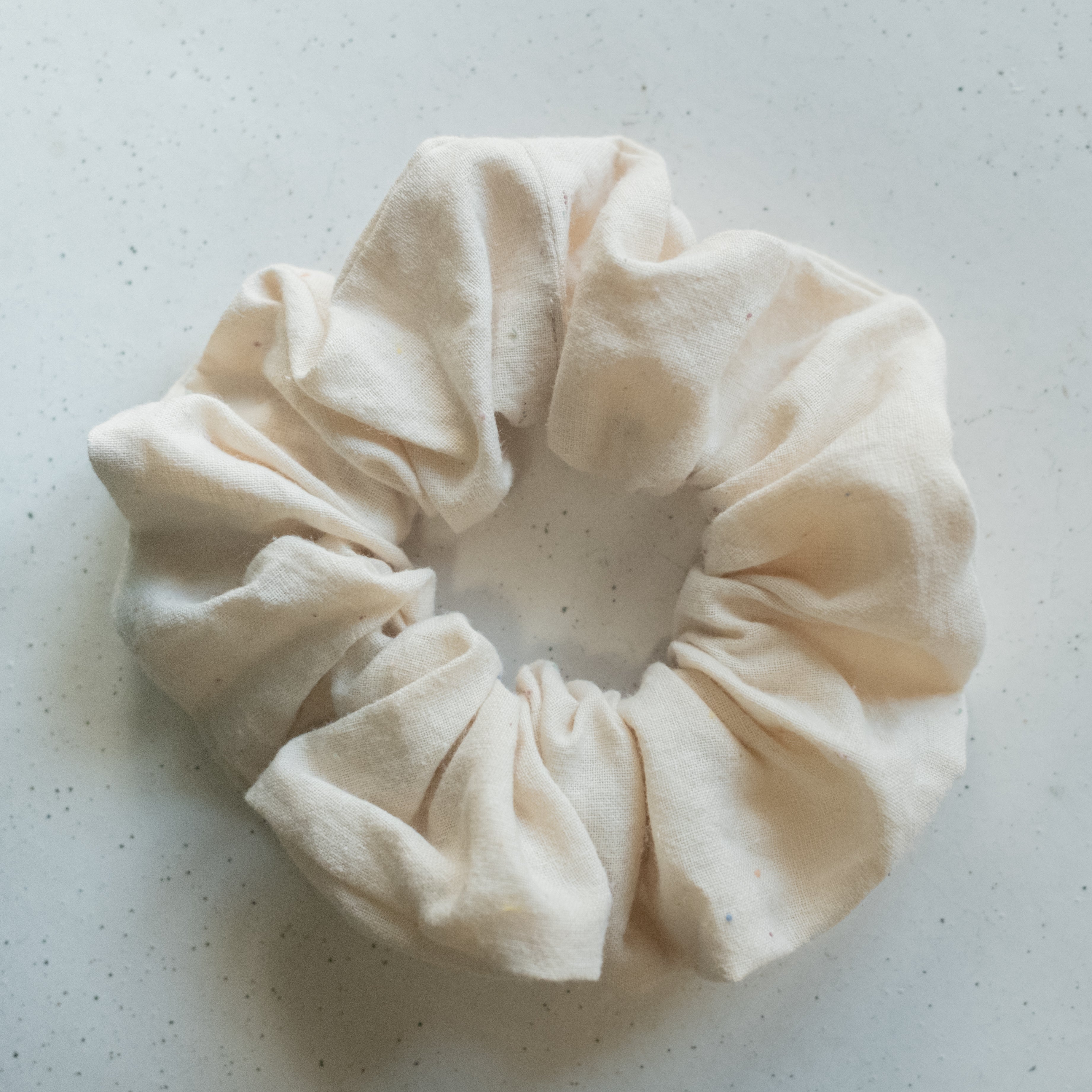A single oversized scrunchie in natural white woven fabric with tiny scattered confetti-coloured specks in pink, blue, and red.