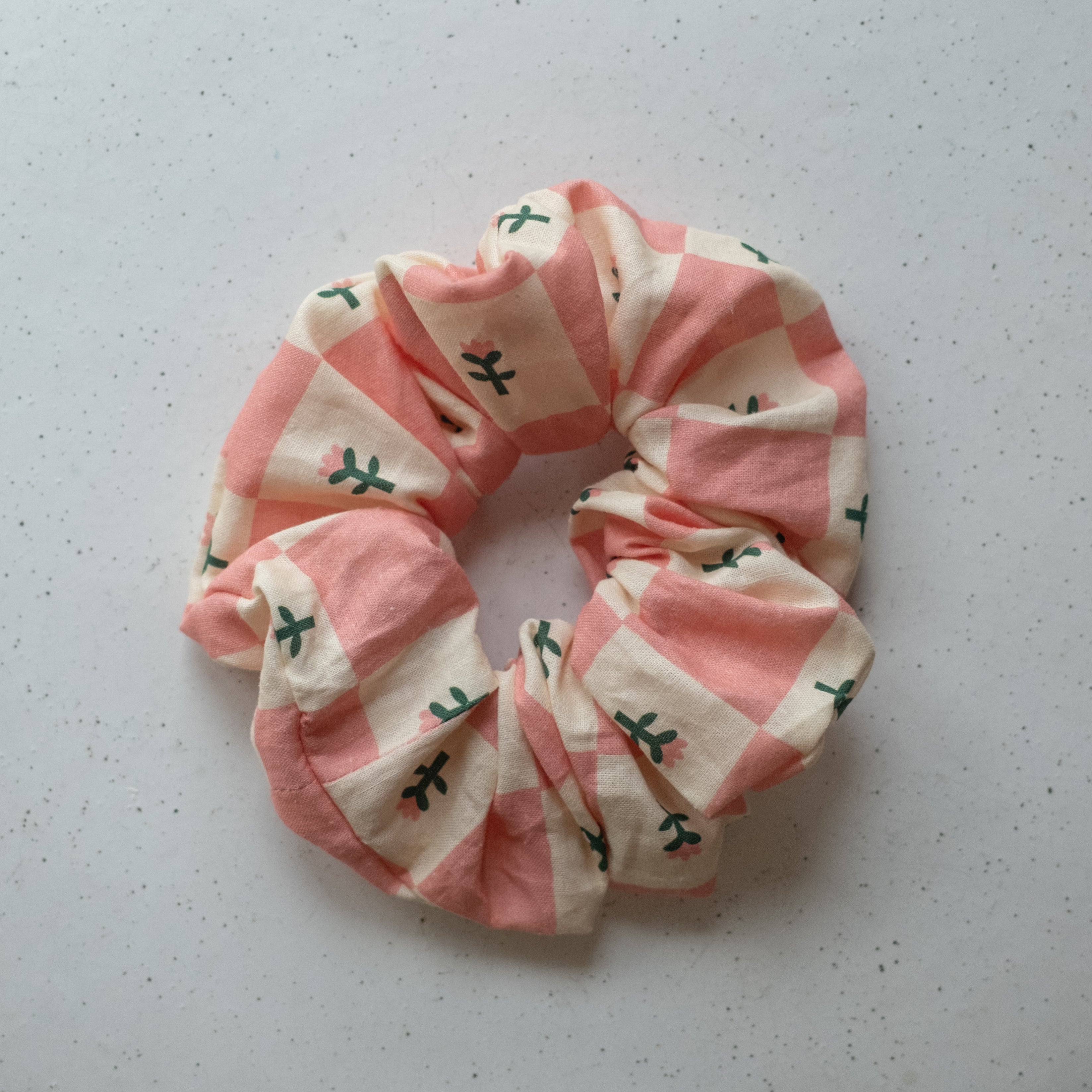 A single oversized scrunchie in pink and cream large-scale checkerboard woven fabric printed with small dark green botanical sprigs and tiny pink flowers.