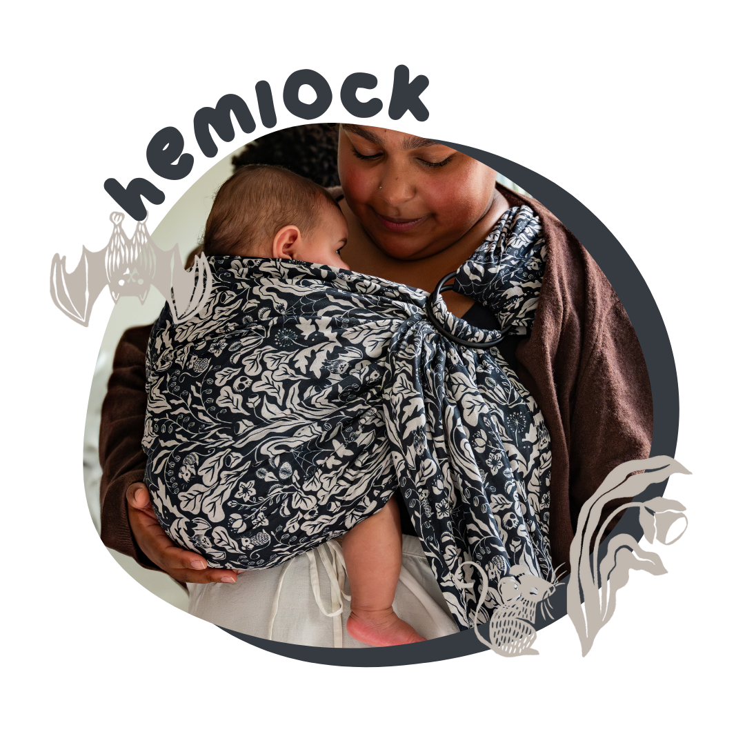 A woman looks tenderly and her baby who is held in a ring sling. The ring sling has a charcoal gray and white print of bats, spiders, mice, and ghosts surrounded by flora.