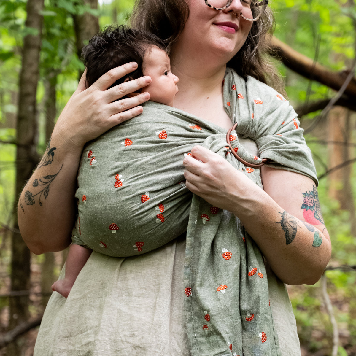 Woman in a wooded setting holding a newborn against her chest in the Amanita Ring Sling, our sage-green chambray fabric, Palm, embroidered with fly agaric red and white spotted mushrooms, with the shoulder ring visible.