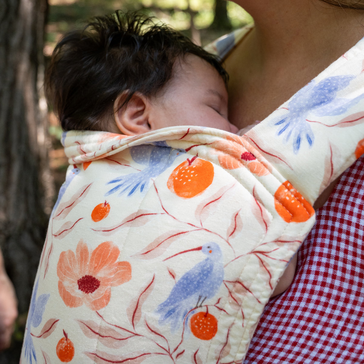 A baby sleeps above the folded headrest of the Ambrosia Lark Baby Carrier, showing blue hummingbirds, coral poppies and orange pomegranates on cream.