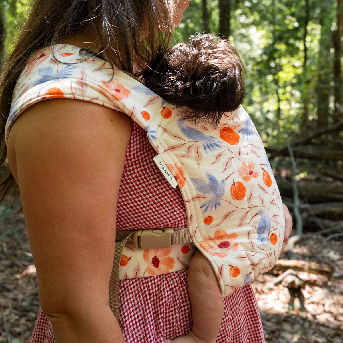 Side view of a baby sleeping in the Ambrosia Lark Baby Carrier, the printed hood and panel supporting them against the wearer's chest.