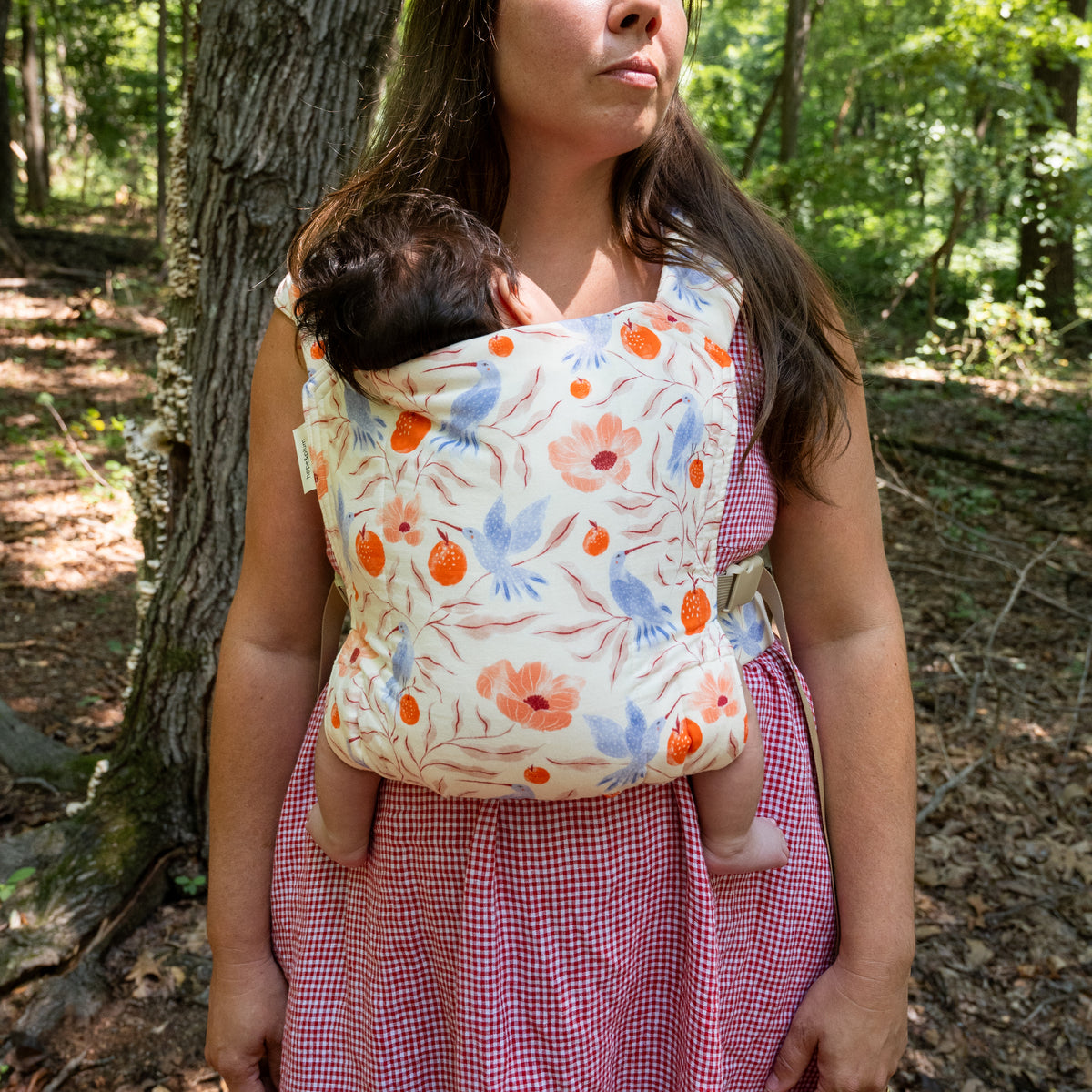 A wearer carries a baby in the Ambrosia Lark Baby Carrier in woodland, the full panel of hummingbirds, coral poppies and pomegranates on cream in view.