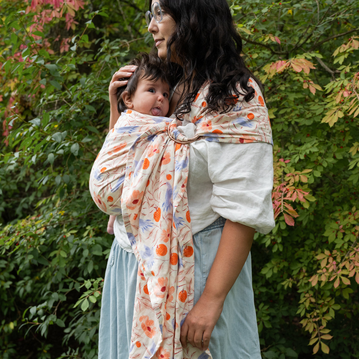 A baby is carried in the Ambrosia Ring Sling outdoors, the rose-gold rings at the shoulder above a print of blue hummingbirds, coral poppies and pomegranates.